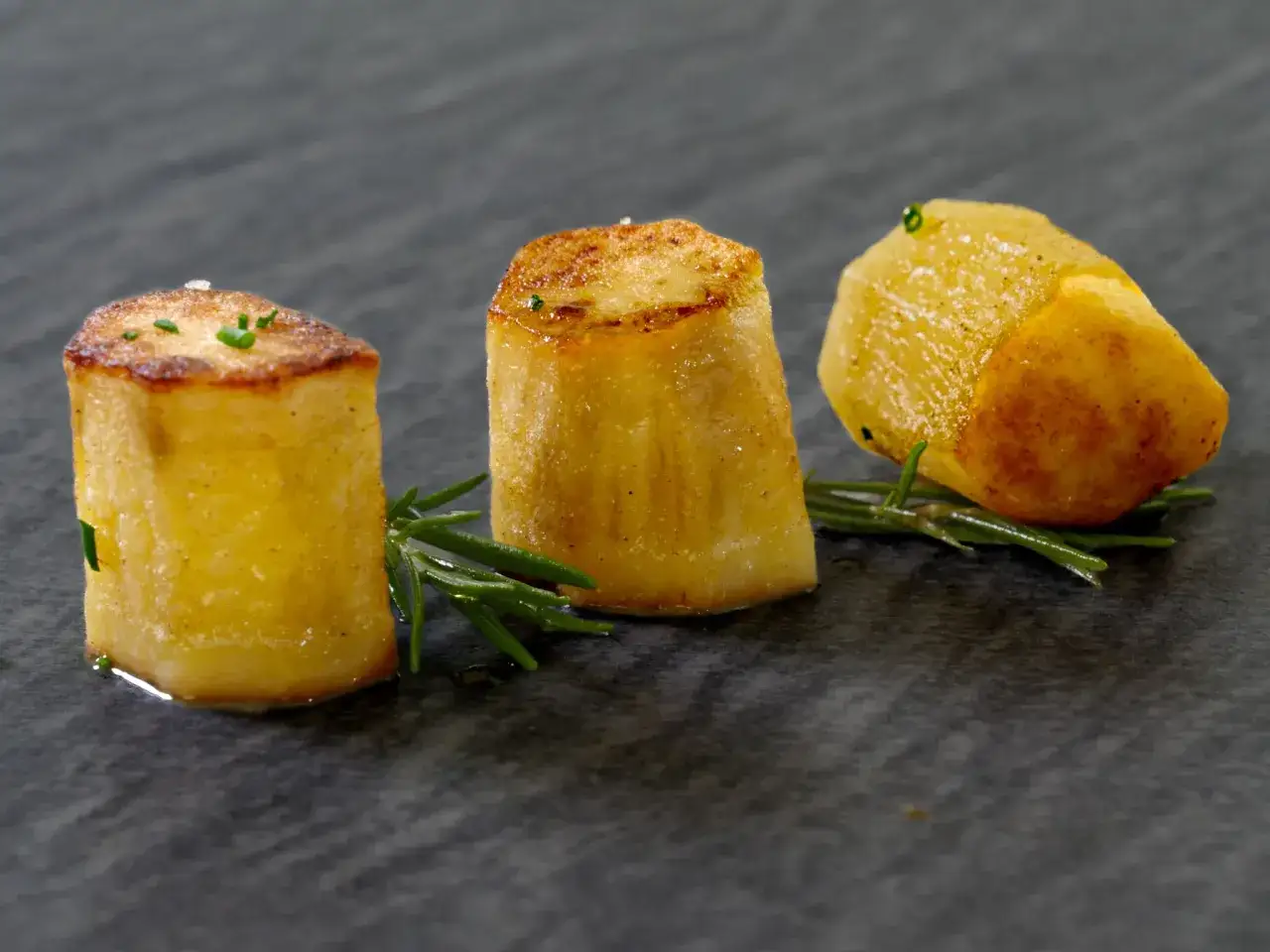 Drei goldbraune Fondantkartoffeln, garniert mit Rosmarin und Schnittlauch, auf dunklem Stein serviert.