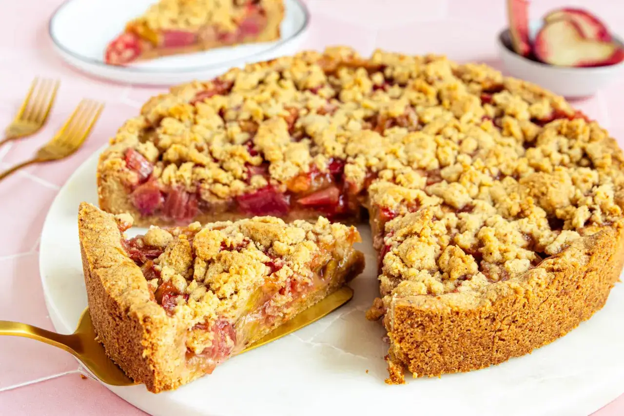 Ein saftiger Rhabarberkuchen in Springform mit knuspriger Streuselhaube. Ein Stück wird gerade herausgehoben.