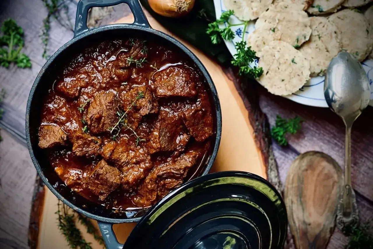 Herzhaftes Gulasch gemischt im Topf, daneben Kl&ouml;&szlig;e und frische Kr&auml;uter. Ein tolles Rezept f&uuml;r gem&uuml;tliche Abende.