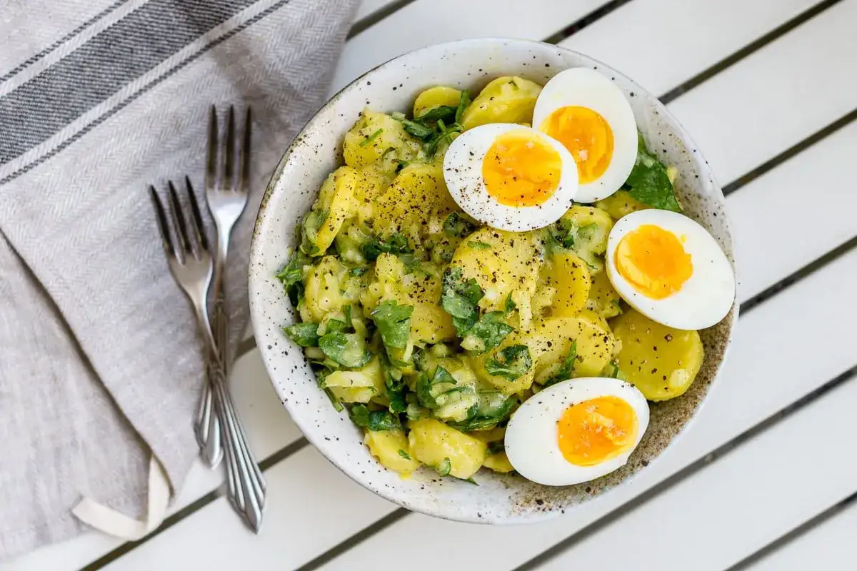 Schüssel mit einfachem Kartoffelsalat, garniert mit gekochten Eiern und frischer Petersilie. Daneben liegen zwei Gabeln auf einer Serviette.