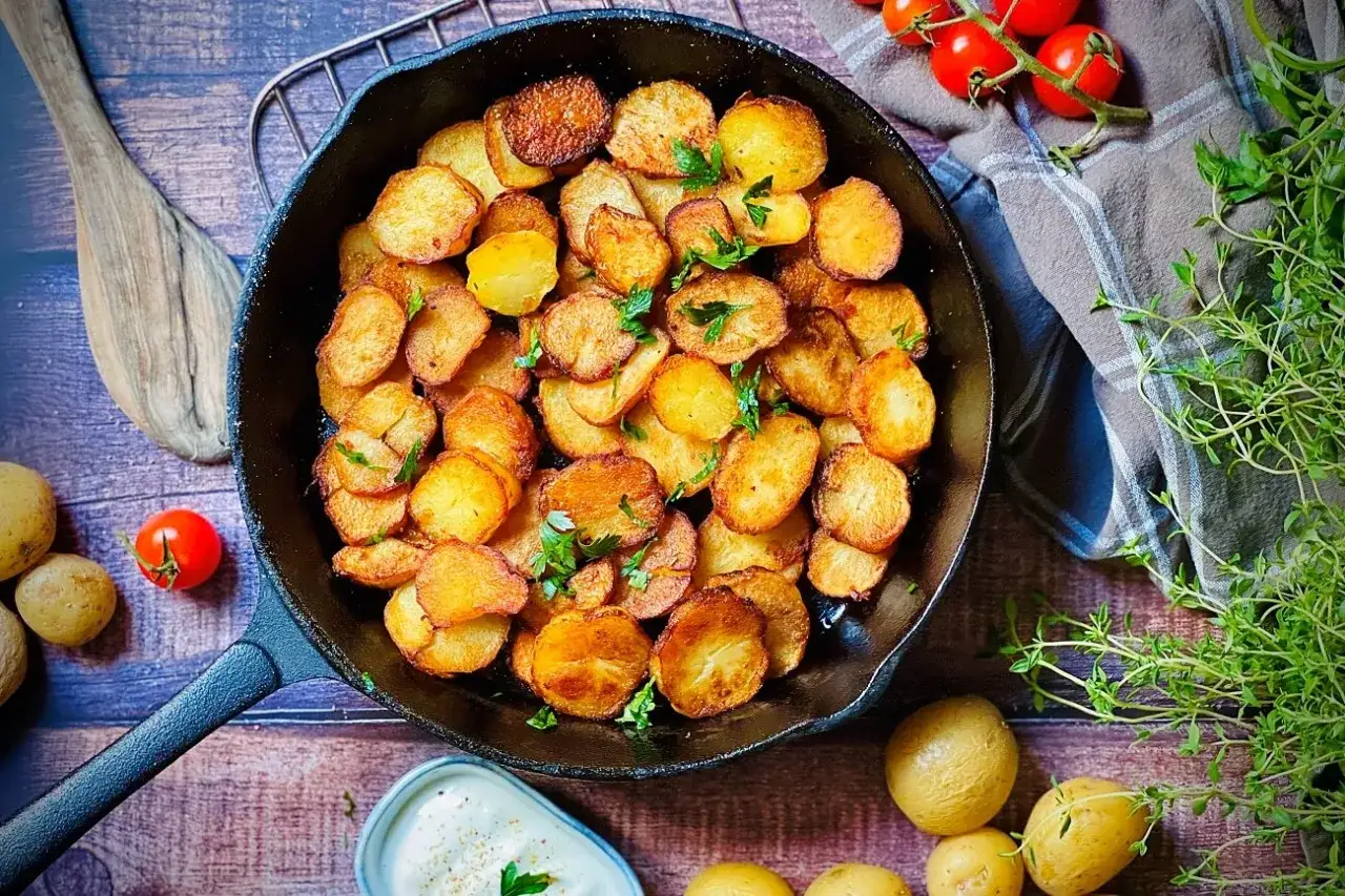 Goldbraun gebratene Kartoffelscheiben in einer Gusseisenpfanne, garniert mit Petersilie. Dazu frische Tomaten und Kräuter. Perfekt zum Kartoffel braten.