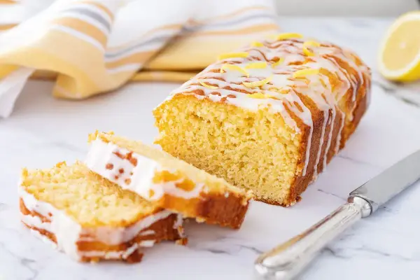 Saftiger zitronenkuchen mit Joghurt, überzogen mit Zuckerguss und garniert mit Zitronenschale. Ein Stück ist angeschnitten.