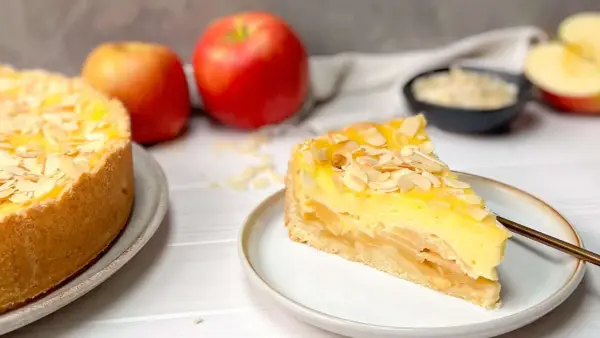 Ein saftiger Apfelkuchen mit Schmandguss, bestreut mit Mandelblättchen. Daneben liegen frische Äpfel.