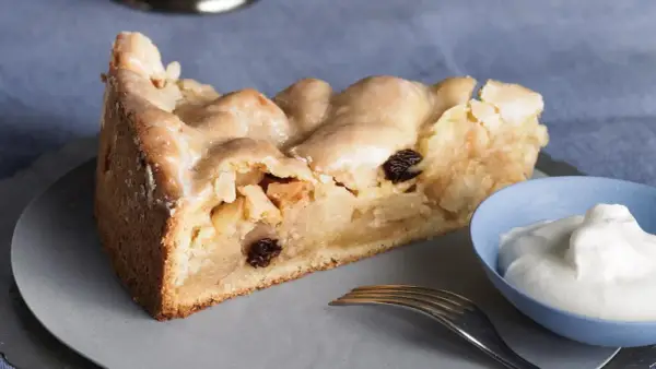 Ein Stück gedeckter Apfelkuchen mit Rosinen und Zuckerguss, serviert mit einem Klecks Sahne.