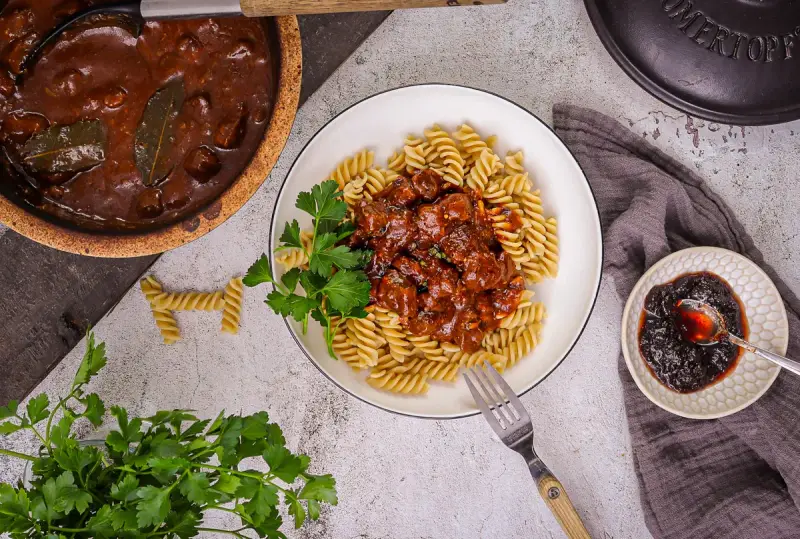 Herzhaftes Gulasch gemischt Rezept mit Fusilli, garniert mit Petersilie. Daneben ein kleiner Teller mit Preiselbeeren.