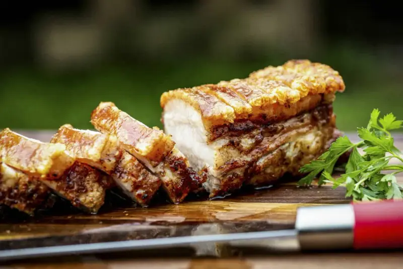 Knuspriger Schweinebauch, perfekt für ein köstliches schweinebauch rezept. Saftig, mit krosser Schwarte, auf einem Holzbrett mit Petersilie und einem Messer.